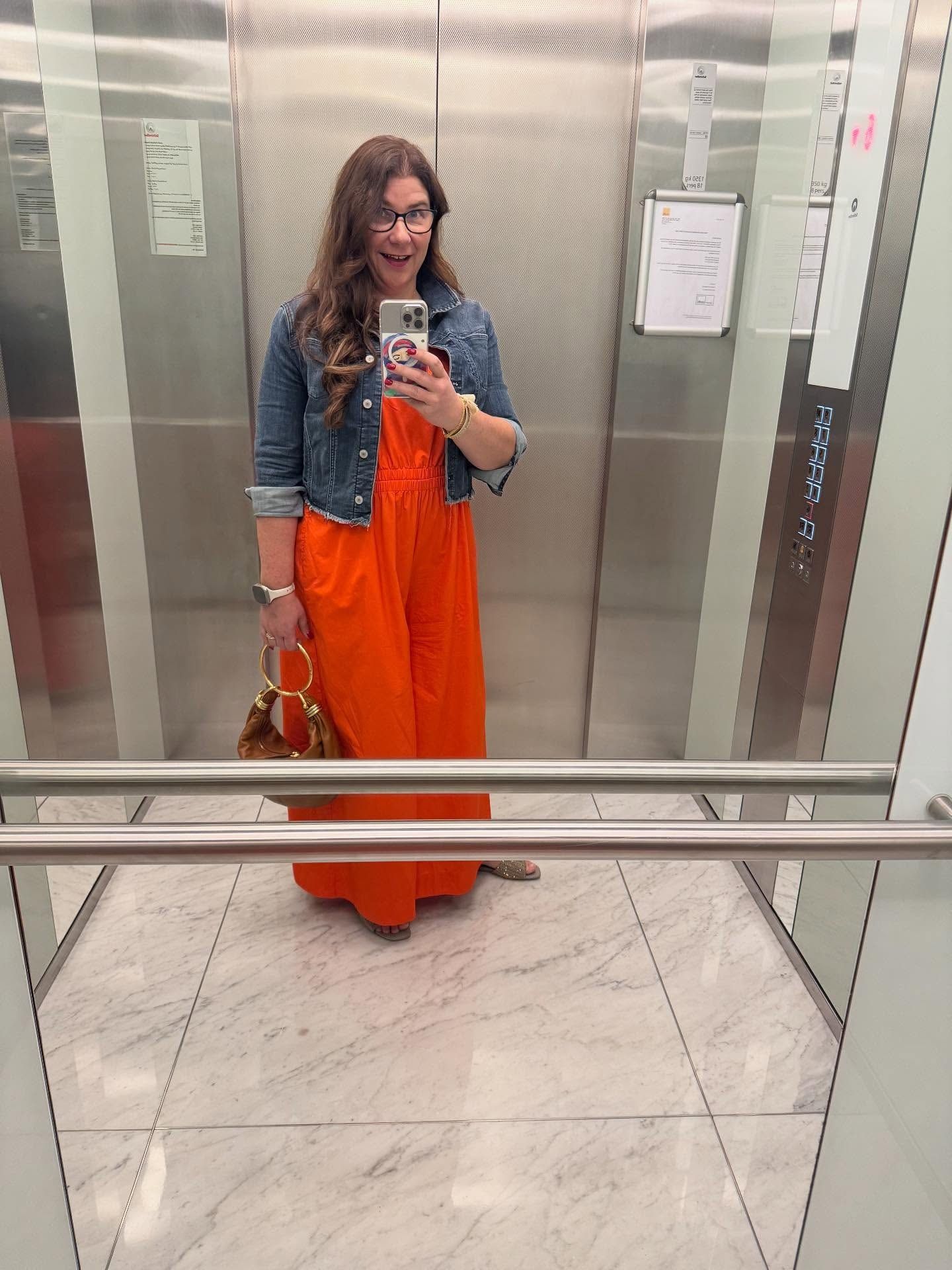 Vanessa in an orange jumpsuit and denim jacket, mirror selfie in an elevator
